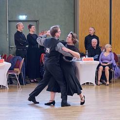 Tanzendes Paar in eleganter Kleidung auf einer Tanzfl&auml;che, umgeben von Zuschauern und Tischen.