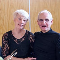 Ein Paar sitzt nebeneinander und l&auml;chelt in die Kamera, vor einem hellen Holzhintergrund.