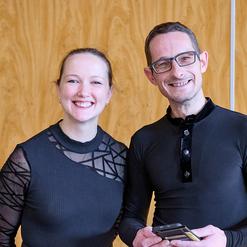 L&auml;chelndes Paar steht vor einer Holzpaneelen-Wand, beide in schwarzen Oberteilen, der Mann h&auml;lt ein Smartphone.