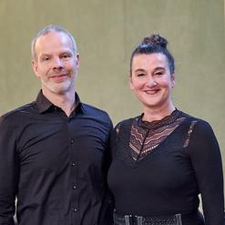 Portr&auml;t von einem Mann in schwarzem Hemd und einer Frau in schwarzem Oberteil mit Spitze, beide l&auml;cheln freundlich.