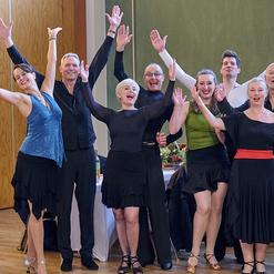 Eine Gruppe von acht tanzenden Personen in eleganter Kleidung hebt die Arme in die Luft und l&auml;chelt fr&ouml;hlich.