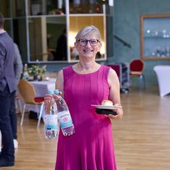 Frau in pinkem Kleid h&auml;lt zwei Wasserflaschen und einen Teller mit einem St&uuml;ck Kuchen in einem festlichen Raum.