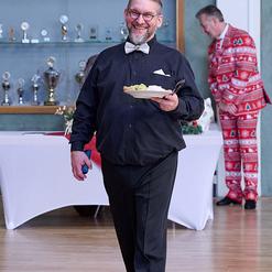 L&auml;chelnder Mann in schwarzem Hemd und Fliege tr&auml;gt einen Teller mit Essen, w&auml;hrend ein anderer Mann im Hintergrund steht.