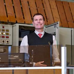 L&auml;chelnder Mann in Weste hinter einem Empfangstresen mit Technik und Steuerungselementen im Hintergrund.