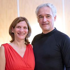 Portrait eines l&auml;chelnden Paares, sie in rot, er in schwarz, vor einer hellen Holzoberfl&auml;che.