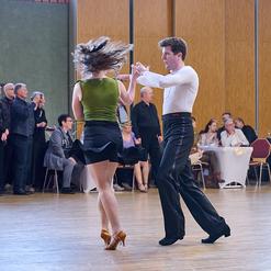 Tanzpaar in Bewegung auf der Tanzfl&auml;che, Zuschauer im Hintergrund, festliche Atmosph&auml;re in einem Saal.