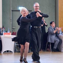 Tanzendes Paar in eleganter Kleidung, umgeben von applaudierendem Publikum in einem Saal.