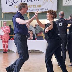 Tanzpaar in formeller Kleidung tanzt fr&ouml;hlich in einem Sportclub, im Hintergrund Zuschauer und ein Banner des Clubs.