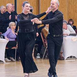 Tanzpaar in eleganter Kleidung beim Tanzen auf einer Veranstaltung, Zuschauer beobachten im Hintergrund.