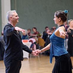 Tanzpaar in eleganter Kleidung beim Gesellschaftstanz in einem festlichen Saal mit anderen T&auml;nzern im Hintergrund.