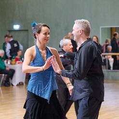 T&auml;nzerpaar in festlicher Kleidung beim Gesellschaftstanz in einem Saal mit mehreren Zuschauern im Hintergrund.