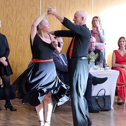 Tanzendes Paar in eleganter Kleidung auf einer Veranstaltung, umgeben von Zuschauern und festlicher Dekoration.