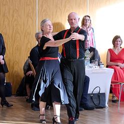 Tanzendes Paar in eleganter Kleidung, umgeben von Zuschauern in festlicher Atmosph&auml;re. Holzfu&szlig;boden im Hintergrund.