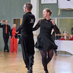 Tanzpaar in eleganter Kleidung w&auml;hrend eines Wettbewerbs auf einem Parkettboden, umgeben von Zuschauern.