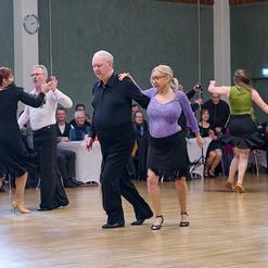 Tanzpaare in eleganter Kleidung tanzen in einem Saal, Zuschauer sitzen an Tischen im Hintergrund.