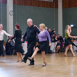 Tanzpaar beim Gesellschaftstanz in formeller Kleidung auf einer hellen Tanzfl&auml;che, im Hintergrund Zuschauer und Tische.