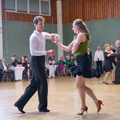Tanzpaar in eleganter Kleidung, das in einem Saal zu Musik tanzt, Zuschauer im Hintergrund.