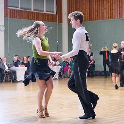 Tanzendes Paar in fr&ouml;hlicher Pose auf einer Tanzfl&auml;che, Zuschauer im Hintergrund beobachten die Darbietung.