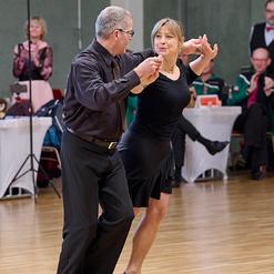 Tanzpaar in eleganter Kleidung tanzt auf einer Holzfl&auml;che, im Hintergrund applaudierende Zuschauer.