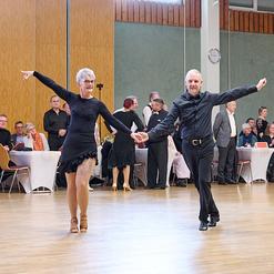 Tanzpaar in eleganter Kleidung f&uuml;hrt einen Tanz auf einer Holzparkettfl&auml;che vor einem Publikum aus mehreren Menschen vor.