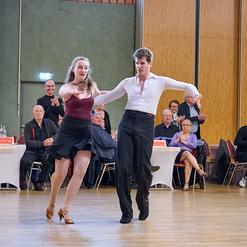 Tanzpaar in eleganter Kleidung f&uuml;hrt einen Tanz auf einer B&uuml;hnenfl&auml;che vor applaudierendem Publikum aus.