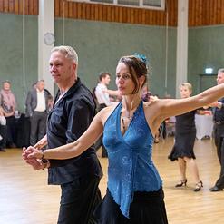 Tanzpaar f&uuml;hrt einen leidenschaftlichen Tanz in einem festlichen Saal mit tanzenden G&auml;sten im Hintergrund aus.
