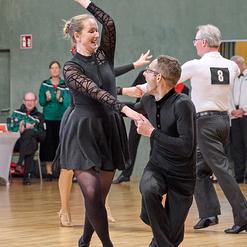 Tanzpaar in formeller Kleidung, w&auml;hrend eines Tanzwettbewerbs auf Holzfl&auml;che, Publikum im Hintergrund.