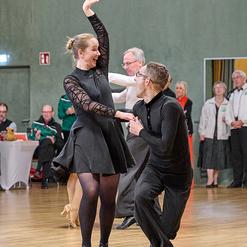 Tanzende Paare in eleganter Kleidung bei einer Veranstaltung in einer h&ouml;lzernen Halle. Die Stimmung ist lebhaft.