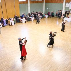 Tanzende Paare in eleganter Kleidung auf einer Holzbodenfl&auml;che, umgeben von Zuschauern und festlicher Dekoration.