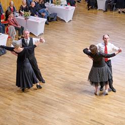 Tanzpaar mit eleganter Kleidung tanzt auf einem Parkettboden, im Hintergrund Zuschauer an Tischen.