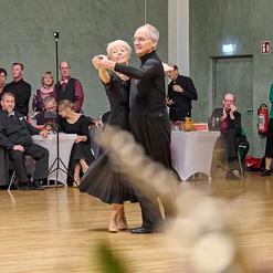 Tanzpaar f&uuml;hrt einen Standardtanz auf einer Tanzfl&auml;che, umgeben von Zuschauern in festlicher Kleidung.