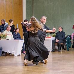 Tanzpaar in eleganter Kleidung f&uuml;hrt einen Tango in einem festlichen Saal mit Zuschauern im Hintergrund auf.