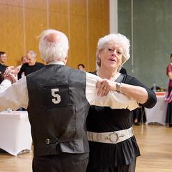 Tanzpaar in eleganter Kleidung bei einer Veranstaltung, umgeben von weiteren T&auml;nzern in einem festlichen Saal.