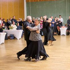 Tanzpaar in eleganter Kleidung tanzt auf einer Tanzfl&auml;che, w&auml;hrend Zuschauer an Tischen im Hintergrund applaudieren.