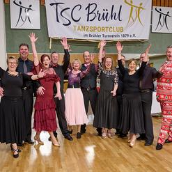Eine Gruppe von T&auml;nzern in formeller Kleidung posiert fr&ouml;hlich vor einem Banner des TSC Br&uuml;hl in einer Tanzhalle.