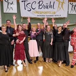 Gruppe von T&auml;nzern in festlicher Kleidung mit erhobenen H&auml;nden vor einem Banner des TSC Br&uuml;hl in einem Tanzsaal.