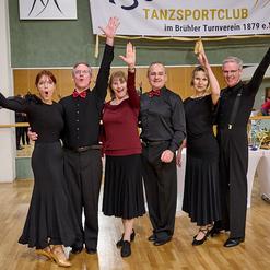 Sechs tanzende Personen in eleganter Kleidung jubeln in einem Tanzsaal mit Spiegelwand und festlicher Dekoration.