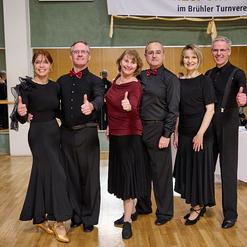Sechs T&auml;nzer in eleganter Kleidung geben Daumen hoch auf einer Tanzveranstaltung, im Hintergrund eine Tanzfl&auml;che.