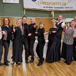 Gruppe von tanzenden Menschen in schwarz, l&auml;chelnd und mit offenen H&auml;nden, in einem Tanzsaal mit Spiegelw&auml;nden.