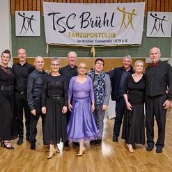 Gruppenfoto von 10 tanzenden Personen in eleganter Kleidung im TSC Br&uuml;hl Tanzenclub mit Banner im Hintergrund.
