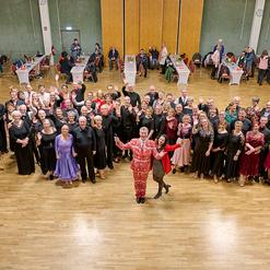 Gruppierung von etwa 60 Personen in eleganter Tanzkleidung, einige winken, in einem festlichen Saal mit Tischen im Hintergrund.