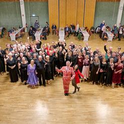 Gruppenszene von 50 tanzenden Personen in festlicher Kleidung, die gemeinsam in einem Saal posieren und l&auml;cheln.