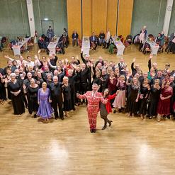 Gruppenaufnahme von Tanzenden in eleganter Kleidung, die jubelnd in einem festlich dekorierten Saal stehen.