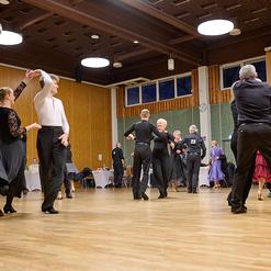 Tanzende Paare in eleganter Kleidung auf einer Holzfl&auml;che, umgeben von Tischen in einem festlichen Saal.