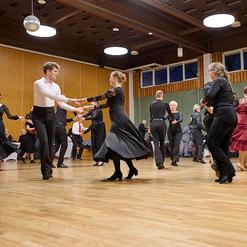 Tanzende Paare in schwarz und rot auf einer Holztanzfl&auml;che, umgeben von Zuschauern in einem festlichen Saal.