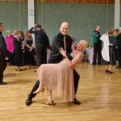 Tanzendes Paar in eleganter Kleidung, umgeben von G&auml;sten auf einer Tanzfl&auml;che in einem festlichen Saal.