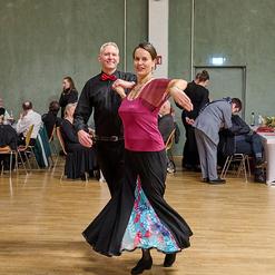Tanzendes Paar in eleganter Kleidung, umgeben von G&auml;sten in einem festlichen Saal mit Holzw&auml;nden.
