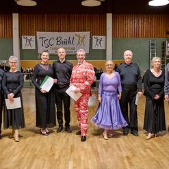 Gruppe von T&auml;nzern in festlicher Kleidung steht auf einer Tanzfl&auml;che in einem Sportclub, im Hintergrund eine Veranstaltungsgemeinschaft.