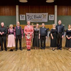 Gruppenfoto von T&auml;nzern in formeller Kleidung vor einer Wand mit dem Logo des TSC Br&uuml;hl in einem Tanzsaal.