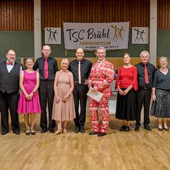 Gruppe von Tanzsportlern in formeller Kleidung, posierend vor einem Banner des TSC Br&uuml;hl im Hintergrund.